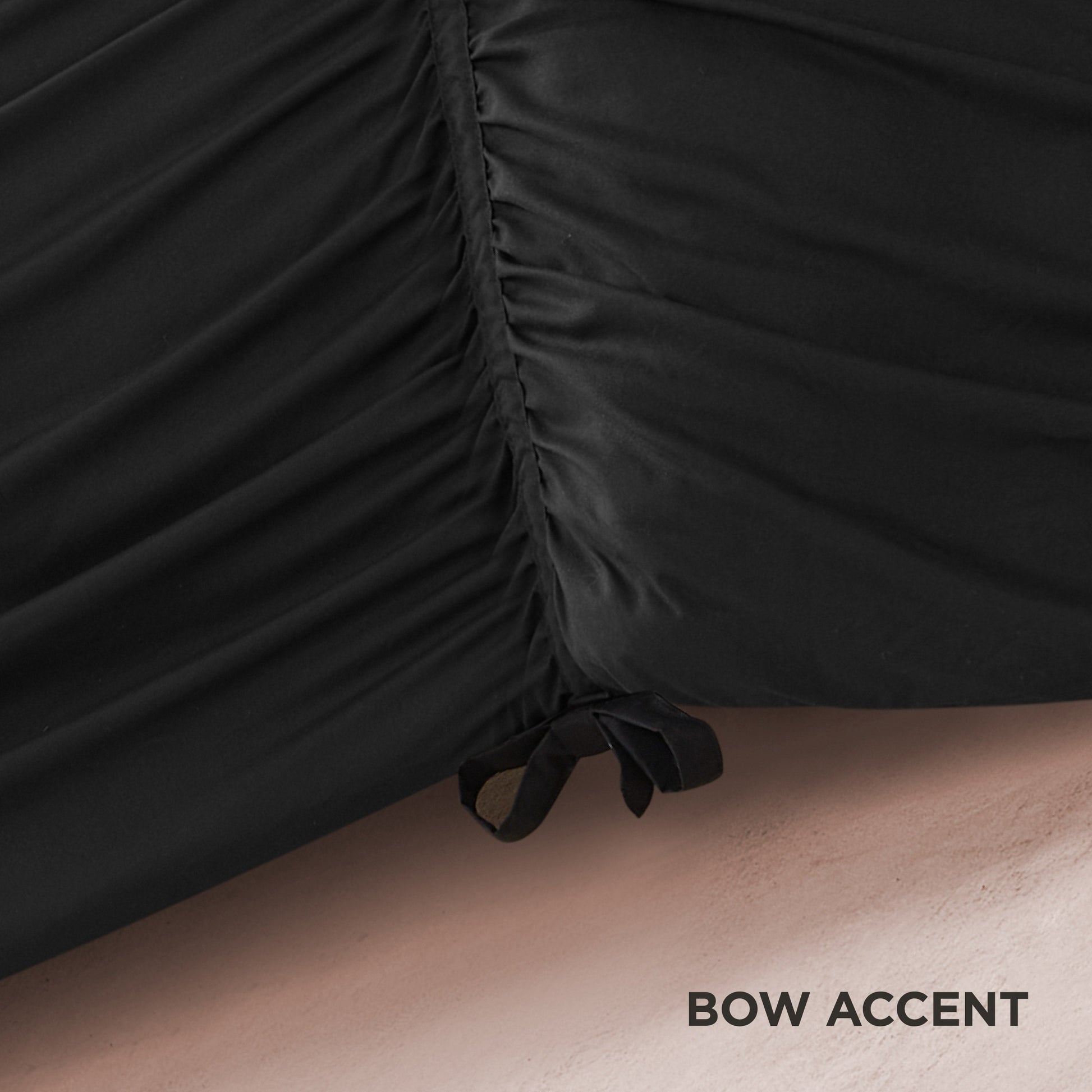 Close-up of black ruched fabric from the Ribbon Ruched Comforter Set, featuring a gathered seam and a delicate ribbon bow at the bottom right. The textured background displays “BOW ACCENT” in the lower right corner.