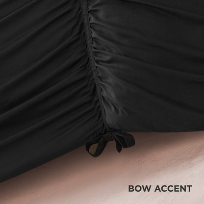 Close-up of black ruched fabric from the Ribbon Ruched Comforter Set, featuring a gathered seam and a delicate ribbon bow at the bottom right. The textured background displays “BOW ACCENT” in the lower right corner.