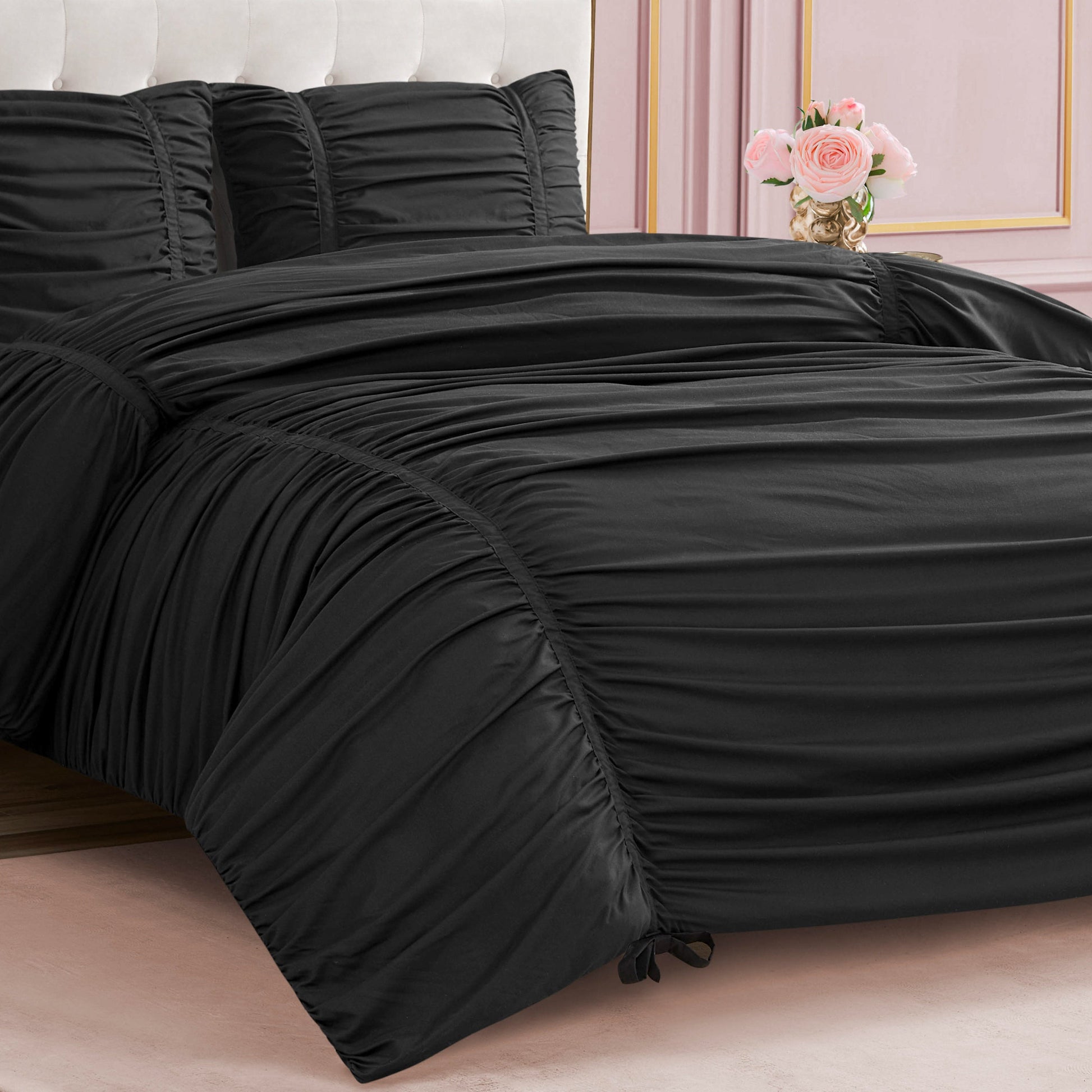 A neatly made bed features the Ribbon Ruched Comforter Set in black with matching pillows, accented by elegant ribbon detailing. Pink walls with decorative molding and a vase of pink roses on the bedside table complete the chic look.