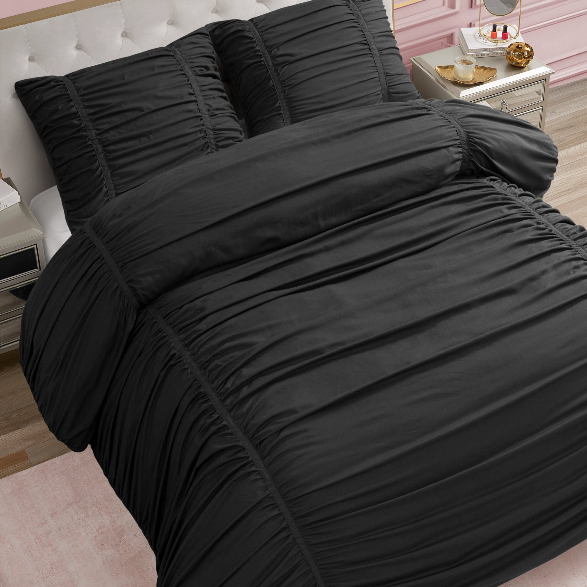 A neatly made bed features the Ribbon Ruched Comforter Set in black with matching pillows, set in a stylish bedroom with a white tufted headboard, pink wall, gold nightstand, and light wood floor.