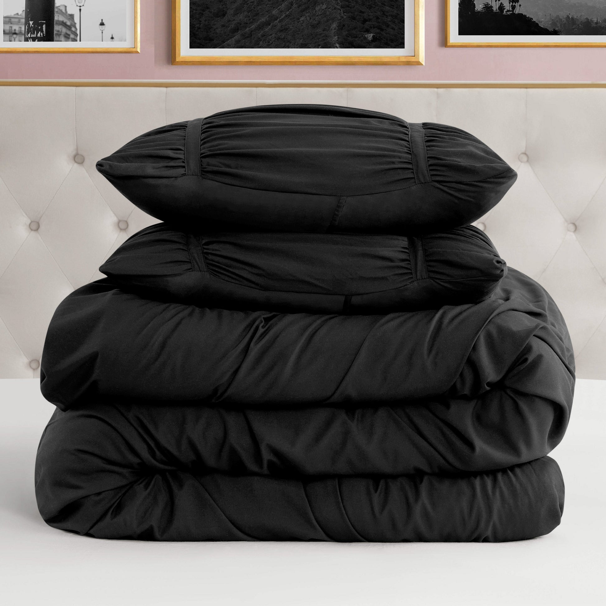 A neatly folded Ribbon Ruched Comforter Set with two matching pillow shams stacked on a white bed, set against a tufted beige headboard and framed pictures on a pink wall.