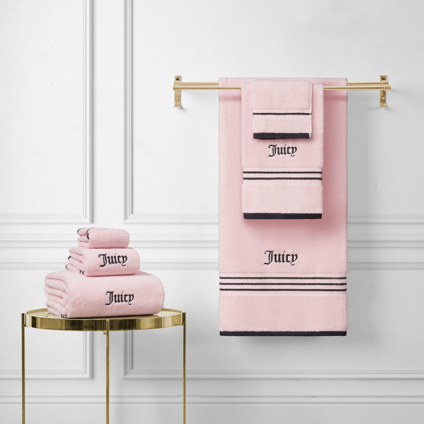 A Neon Border Bath Towel Set in pink with black stripes and "Juicy" embroidery is neatly folded and displayed on a gold towel rack and side table against a white paneled wall.
