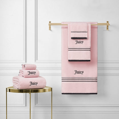 A Neon Border Bath Towel Set in pink with black stripes and "Juicy" embroidery is neatly folded and displayed on a gold towel rack and side table against a white paneled wall.