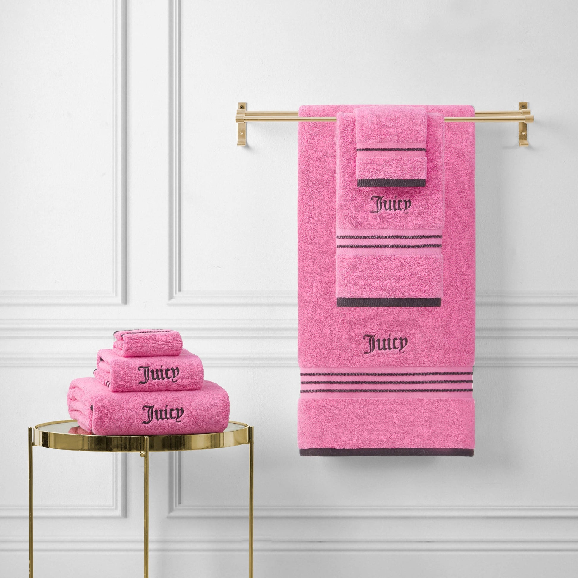 Neon Border Bath Towel Set towels in bright pink, featuring "Juicy" embroidered in black, are neatly folded on a small gold table and displayed on a gold towel rack against a white paneled wall.