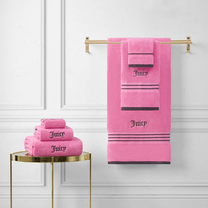 Neon Border Bath Towel Set towels in bright pink, featuring "Juicy" embroidered in black, are neatly folded on a small gold table and displayed on a gold towel rack against a white paneled wall.