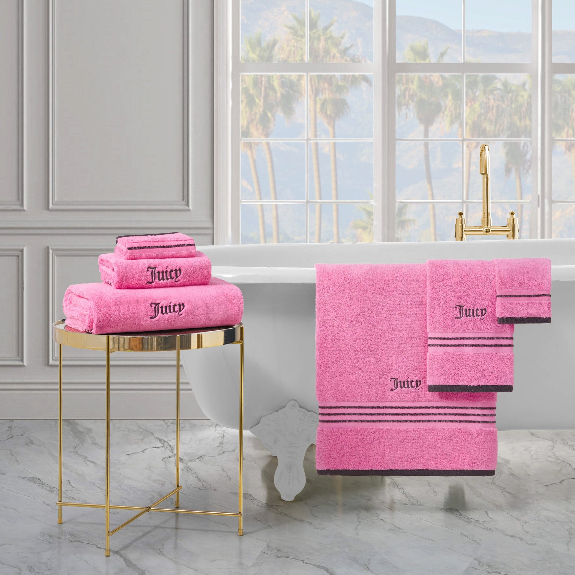 A luxurious bathroom with a white bathtub, gold faucet, and plush Neon Border Bath Towel Set folded on a gold side table and hanging on the tub. Large windows reveal palm trees and mountains outside.