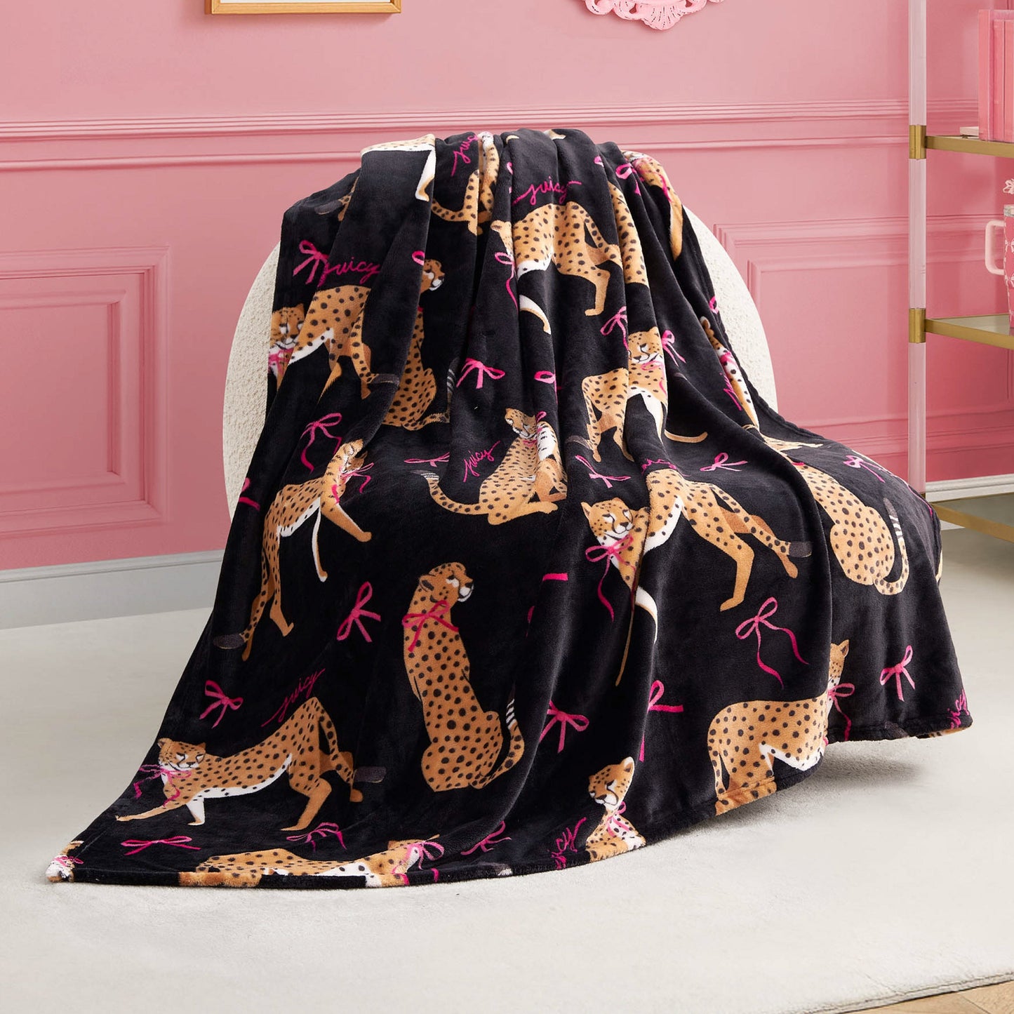The Plush Throw Blanket is a black fleece with a colorful cheetah and pink bow print, shown draped over a white chair in a pink room with decorative wall molding.