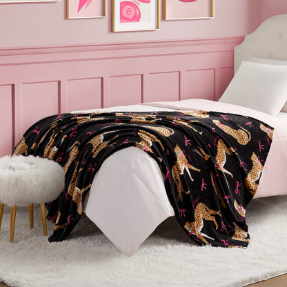 A neatly made bed with a white comforter and a Plush Throw Blanket featuring cheetah and pink bow patterns. The room has pink walls with framed art, a white furry stool, and a white rug on the floor.