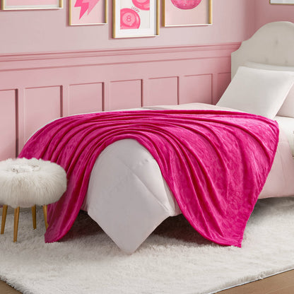 A neatly made bed features a Plush Throw Blanket, bright pink and ultra-soft, draped over white bedding in a pink-themed room. A fur-topped stool and framed wall art complete the cozy, stylish decor.