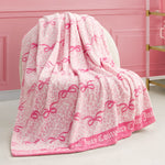 A Plush Throw Blanket featuring pink and white leopard print, pink bow designs, and "Juicy Couture" along the border is draped over a cream-colored chair in a pink room.
