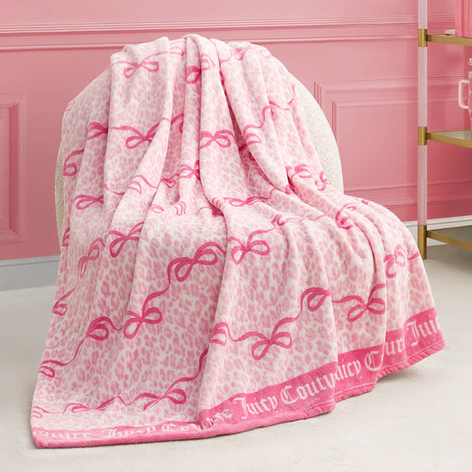 A Plush Throw Blanket featuring pink and white leopard print, pink bow designs, and "Juicy Couture" along the border is draped over a cream-colored chair in a pink room.
