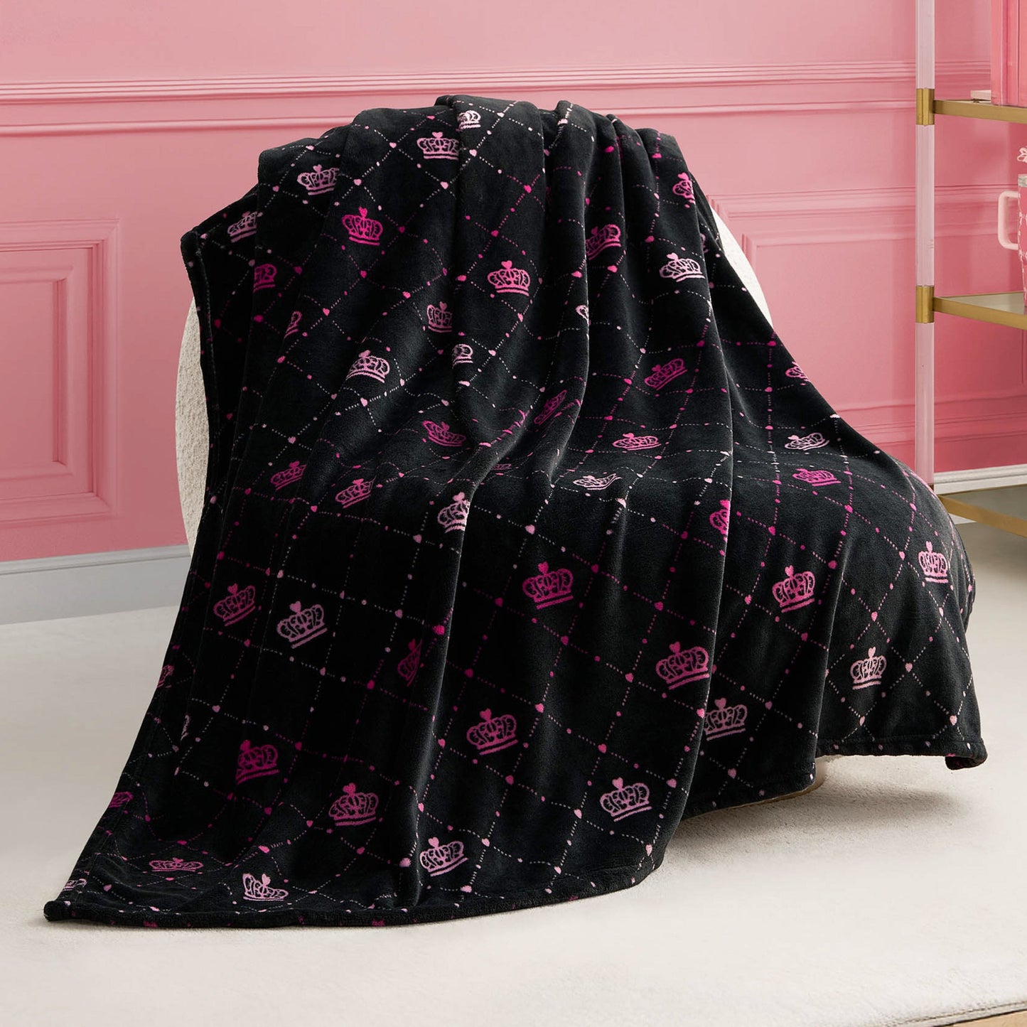 A Plush Throw Blanket in black with a quilted pattern and pink and white crown designs is draped over a white chair in a pink room with elegant wall panels and a glass shelf.