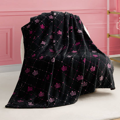 A Plush Throw Blanket in black with a quilted pattern and pink and white crown designs is draped over a white chair in a pink room with elegant wall panels and a glass shelf.