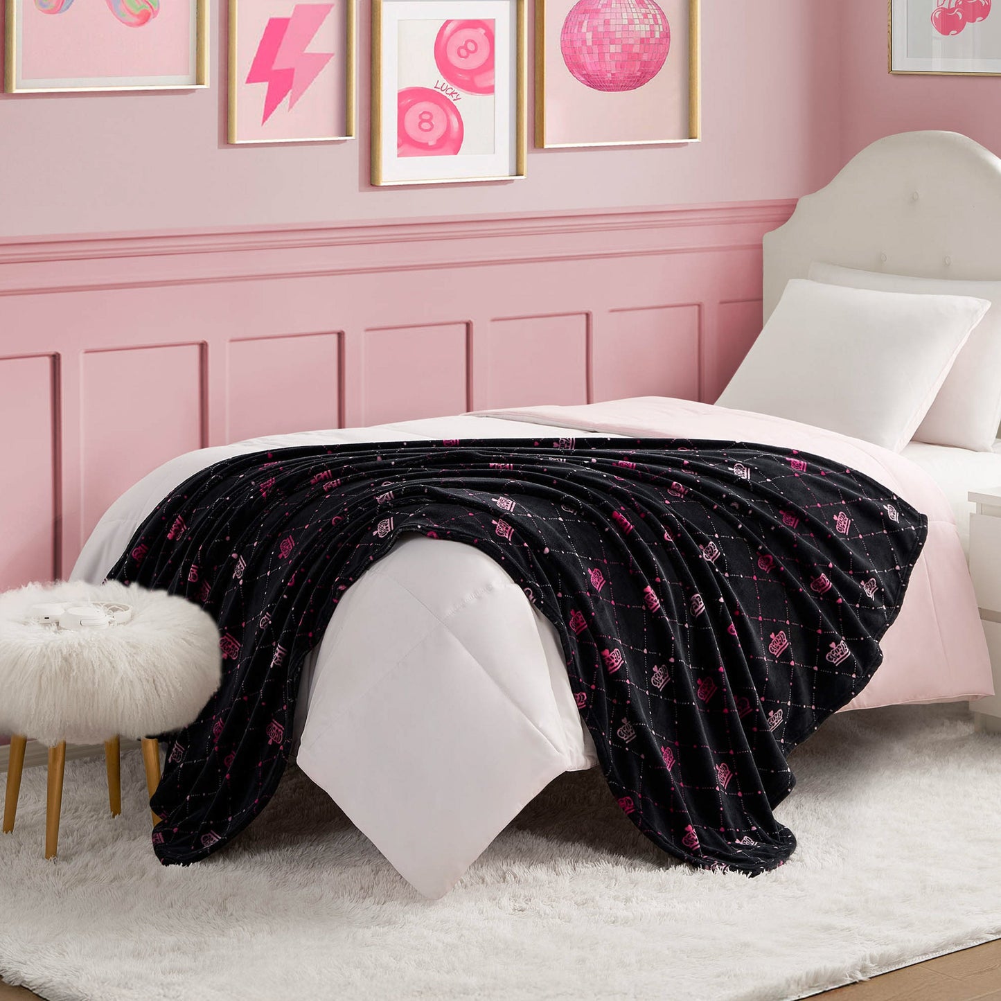 A white bed with a tufted headboard and white bedding features a Plush Throw Blanket with pink patterns. The room has pink walls, framed pink artwork, and a white faux fur stool with wooden legs.