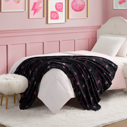 A white bed with a tufted headboard and white bedding features a Plush Throw Blanket with pink patterns. The room has pink walls, framed pink artwork, and a white faux fur stool with wooden legs.