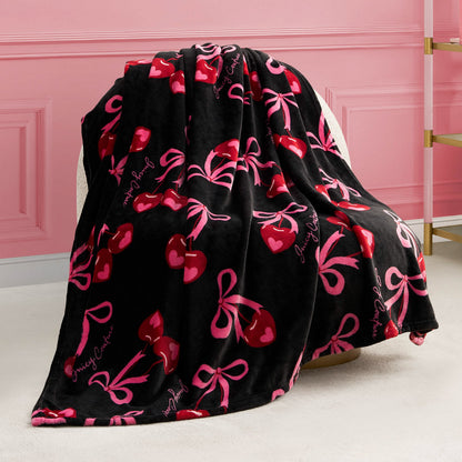The Sweet Cherry Bows Plush Throw Blanket, ultra-soft and black with red cherry and pink bow designs plus pink cursive lettering, is draped over a beige chair against a pink wall with molding.