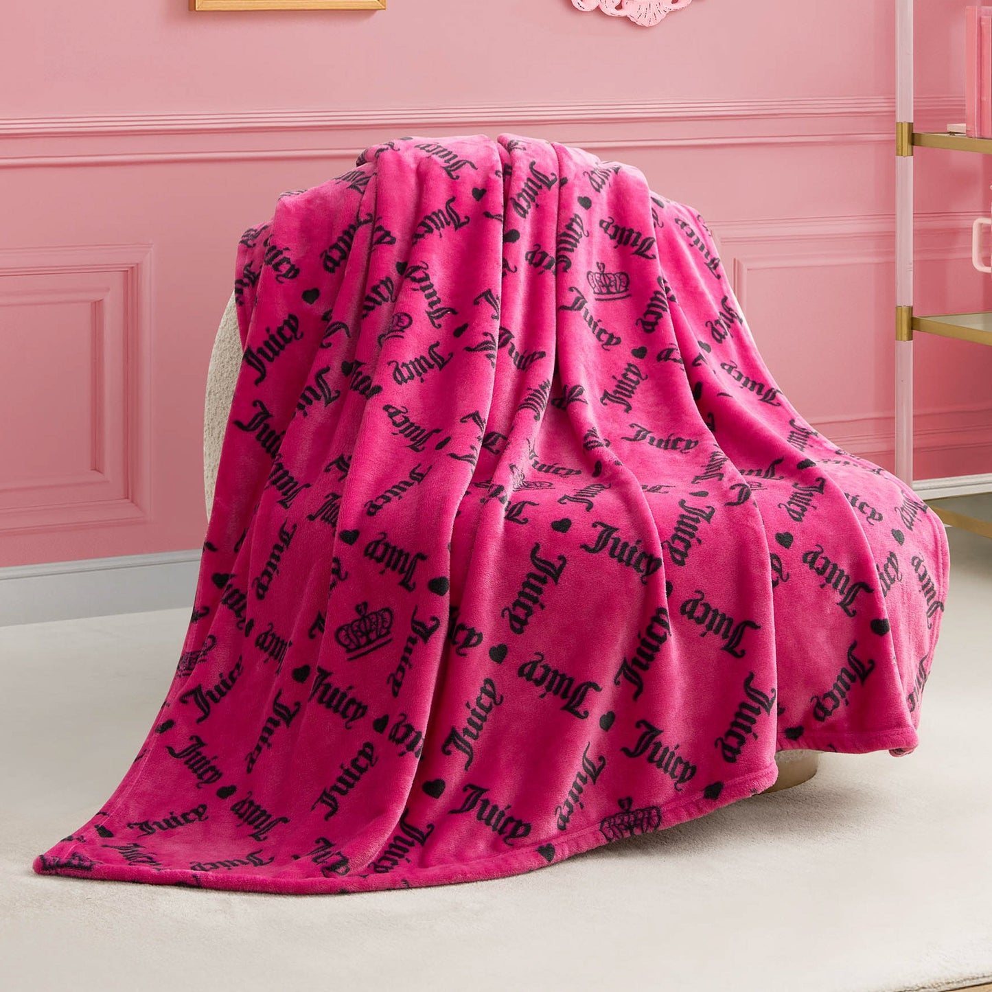 A Plush Throw Blanket in pink, with black Juicy text and crown motifs, is draped over a white chair in a pink room featuring wainscoting and gold-accented shelves for a glamorous vibe.