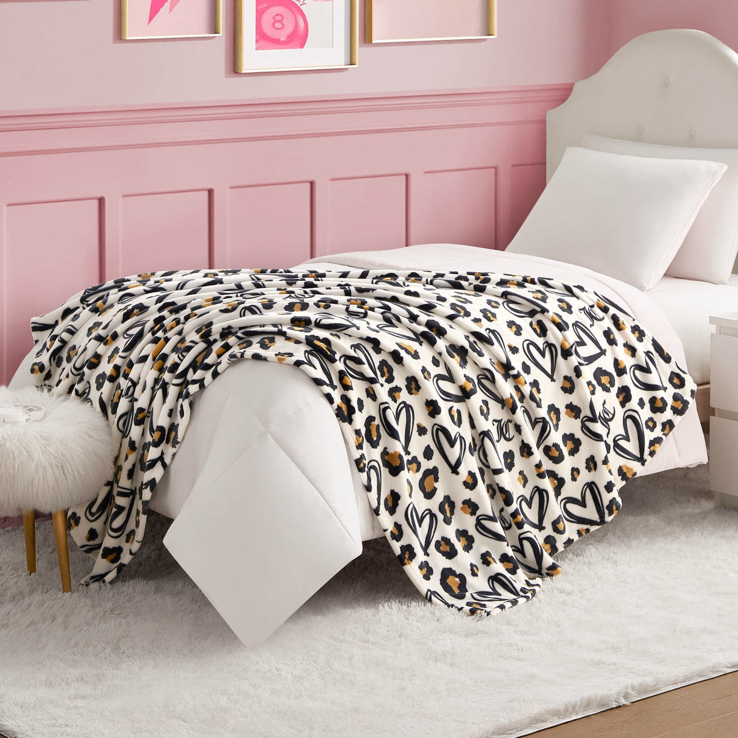 A cozy bed with white bedding and a Plush Throw Blanket featuring black hearts and cheetah spots sits in a pink-walled room with framed art, a white headboard, and a fluffy white rug.