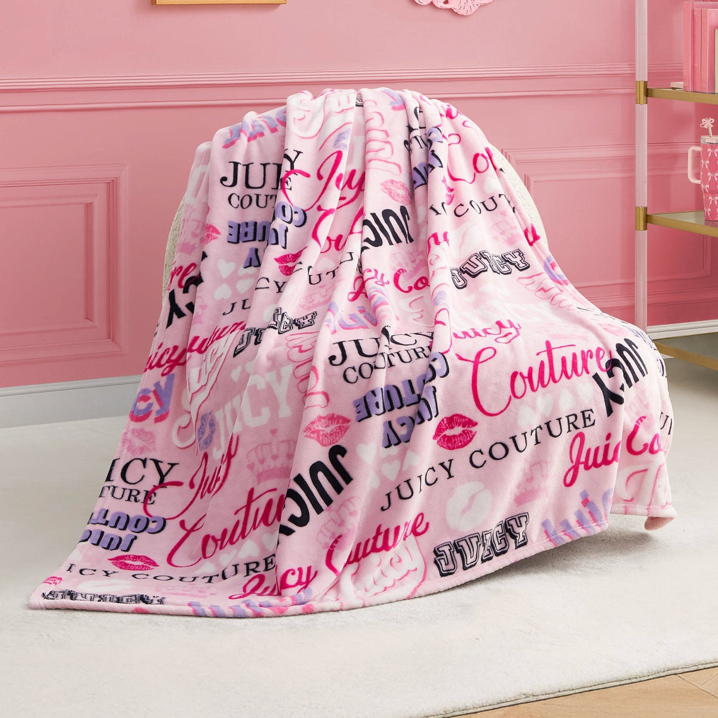 A Plush Throw Blanket with pink and red lip and Juicy Couture logo prints is draped over a white chair in a stylish pink room with gold-accented furniture, adding cozy glamour and ultra-soft comfort to any space.