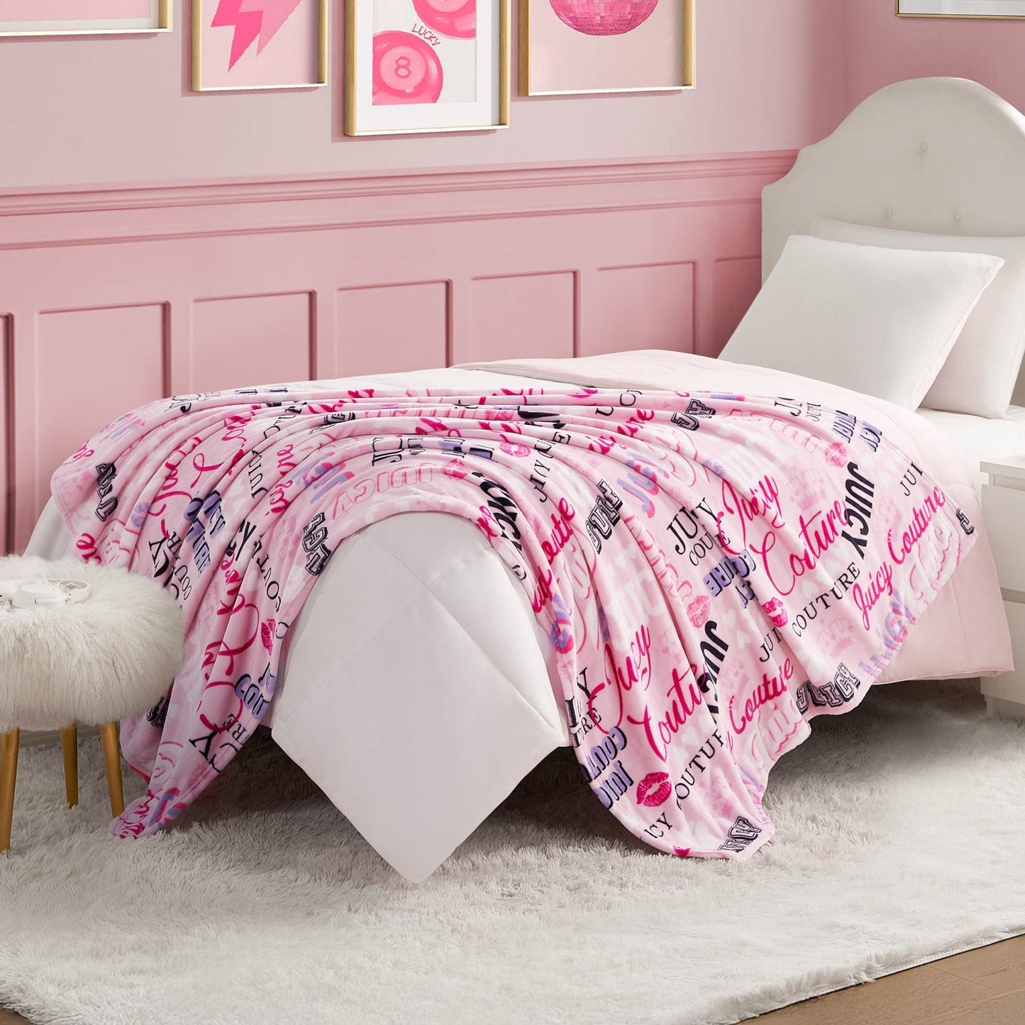 A neatly made bed with a white comforter and a pink Plush Throw Blanket featuring various logo prints, in a pink-themed bedroom with white furniture and framed wall art.