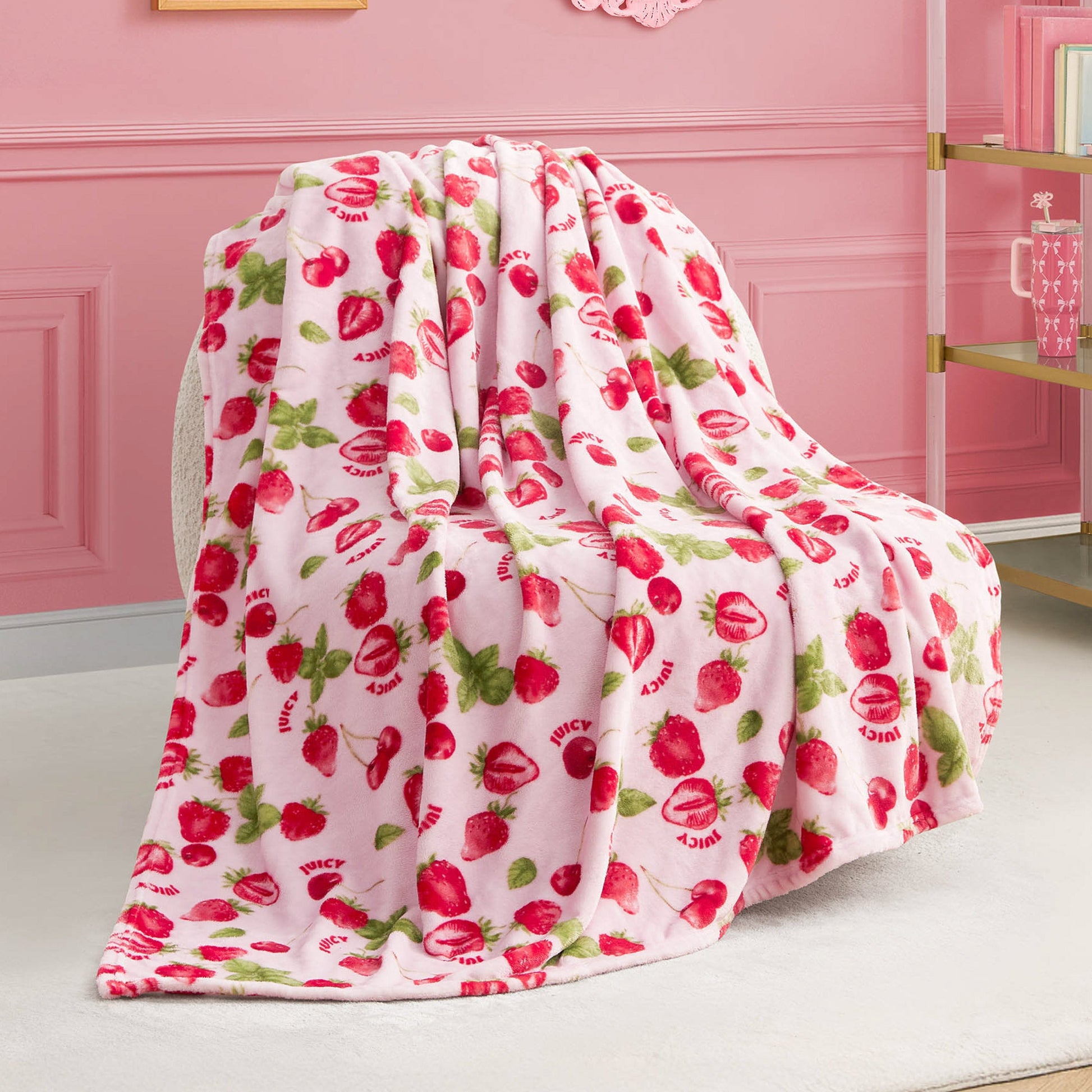 The Plush Throw Blanket features soft pink fleece with red strawberries, green leaves, and “JUICY” lettering—perfect for draping over a chair to accent a pink-themed room with a gold-and-glass shelf in the background.