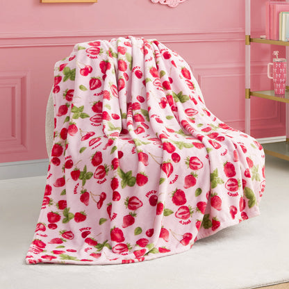 The Plush Throw Blanket features soft pink fleece with red strawberries, green leaves, and “JUICY” lettering—perfect for draping over a chair to accent a pink-themed room with a gold-and-glass shelf in the background.