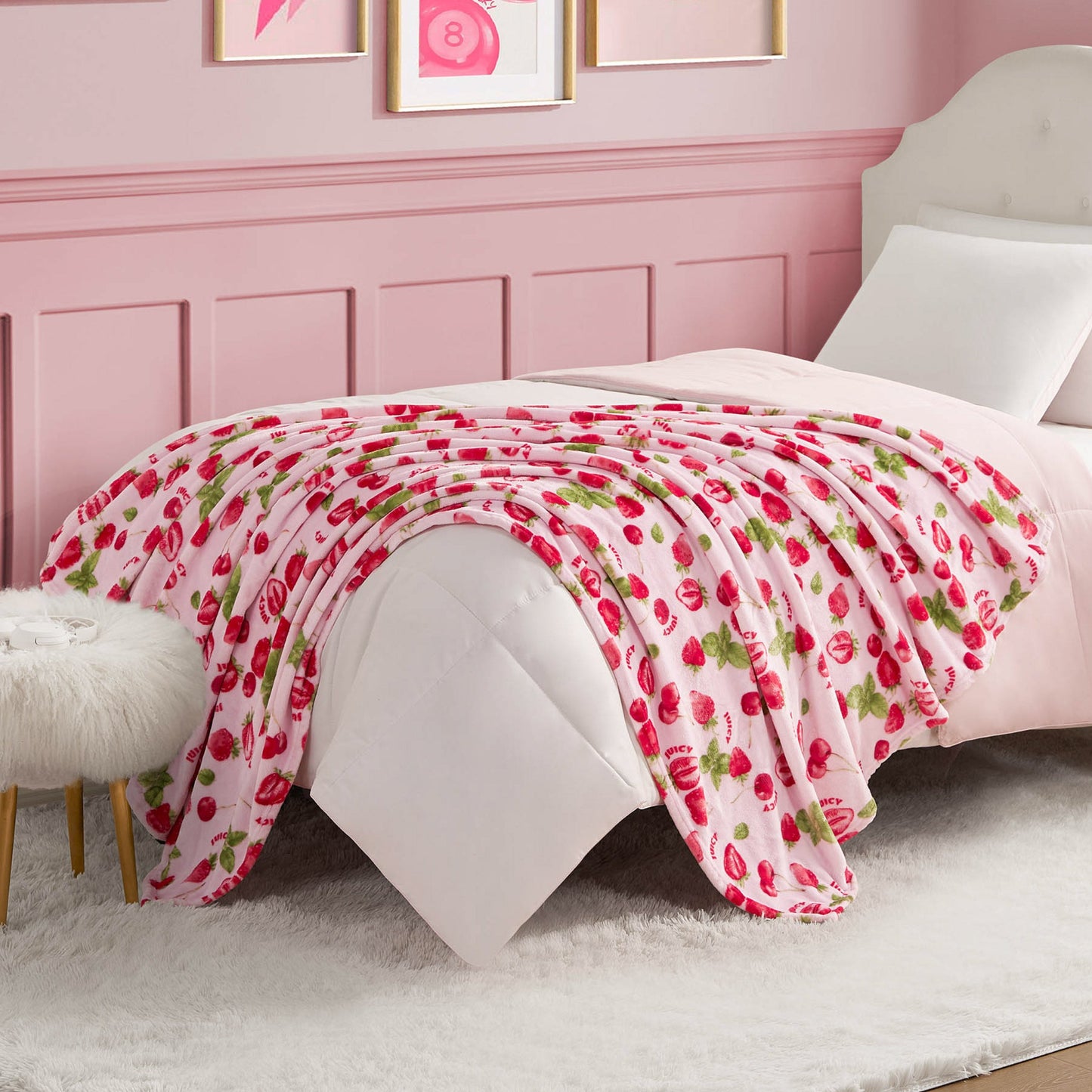 A bed features a white comforter and a pink Plush Throw Blanket decorated with red strawberries, set in a pink-themed bedroom with white and gold accents and wall art.