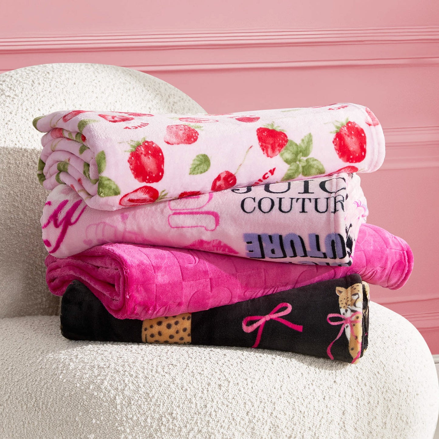 Three folded Plush Throw Blankets sit on a textured white chair: the top is white with strawberry prints, the middle is bright pink with bold text, and the bottom features black leopard spots and pink bow designs.