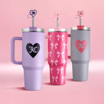 Three Juicy Couture Jetsetter Hydration Stainless Steel Tumblers (40 oz) are shown on a pink background: one lavender with a black heart, one pink with white bows, and one gray with a pink heart. Each includes a straw topped with a heart-shaped decoration.