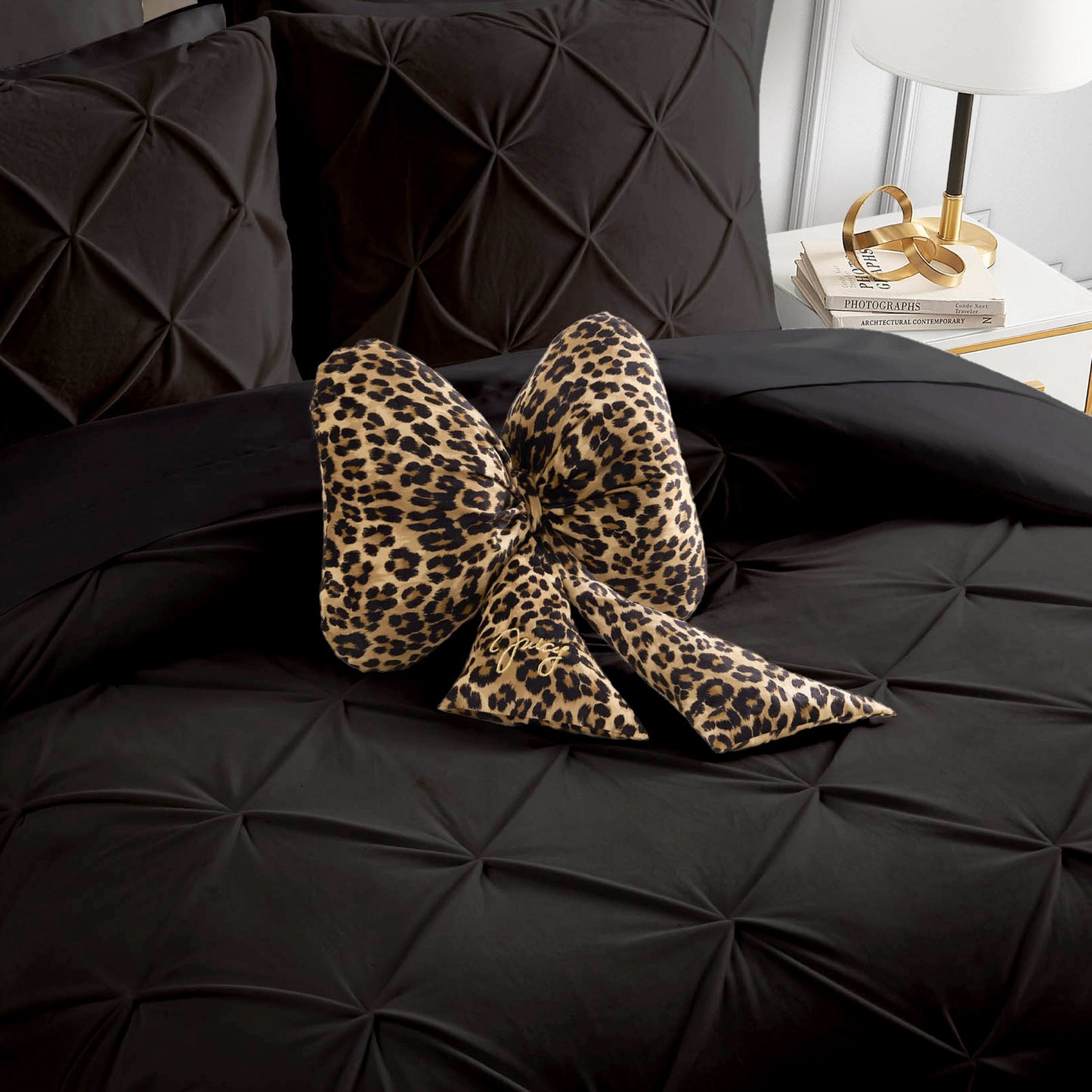 A Bow Shaped Decorative Pillow by Juicy Couture in a bold leopard print rests atop a neatly made black, textured bed. Beside the bed, a white nightstand displays a lamp, several books, and gold accessories.