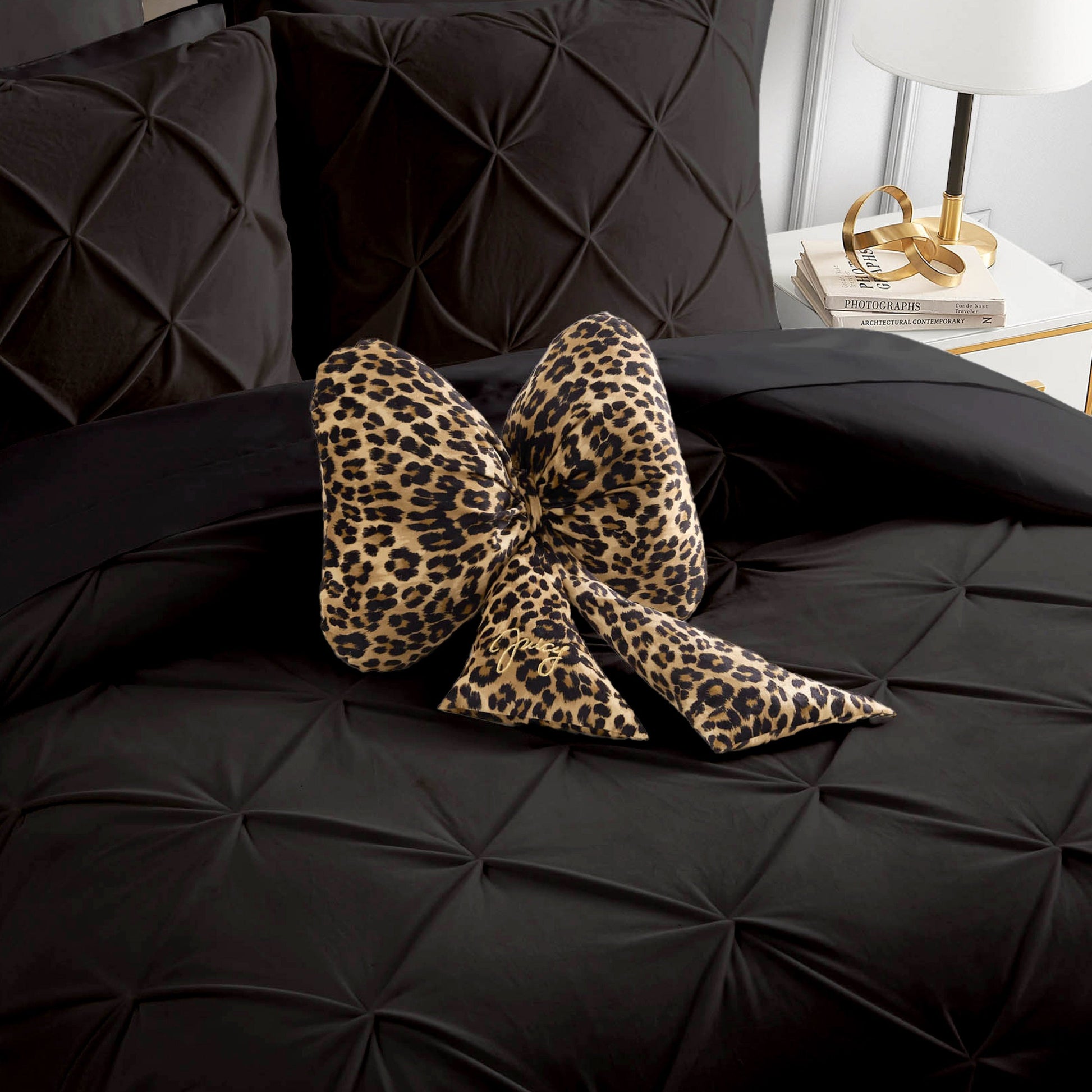 A Bow Shaped Decorative Pillow by Juicy Couture in a bold leopard print rests atop a neatly made black, textured bed. Beside the bed, a white nightstand displays a lamp, several books, and gold accessories.