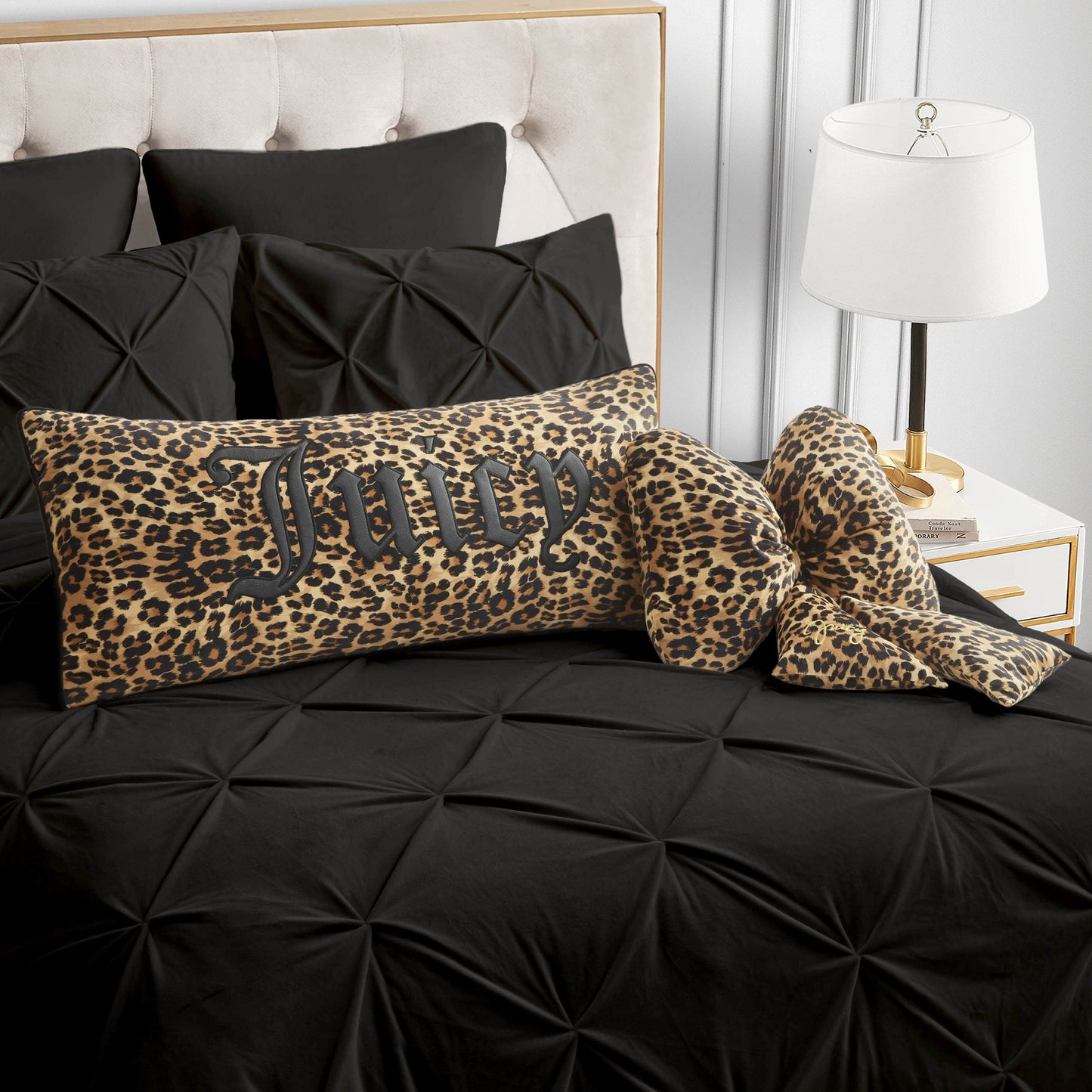 A bed featuring black textured bedding, a Juicy Couture leopard print pillow with "Juicy" written on it, coordinating leopard print slippers, a Bow Shaped Decorative Pillow, and a white lamp placed on the nightstand.