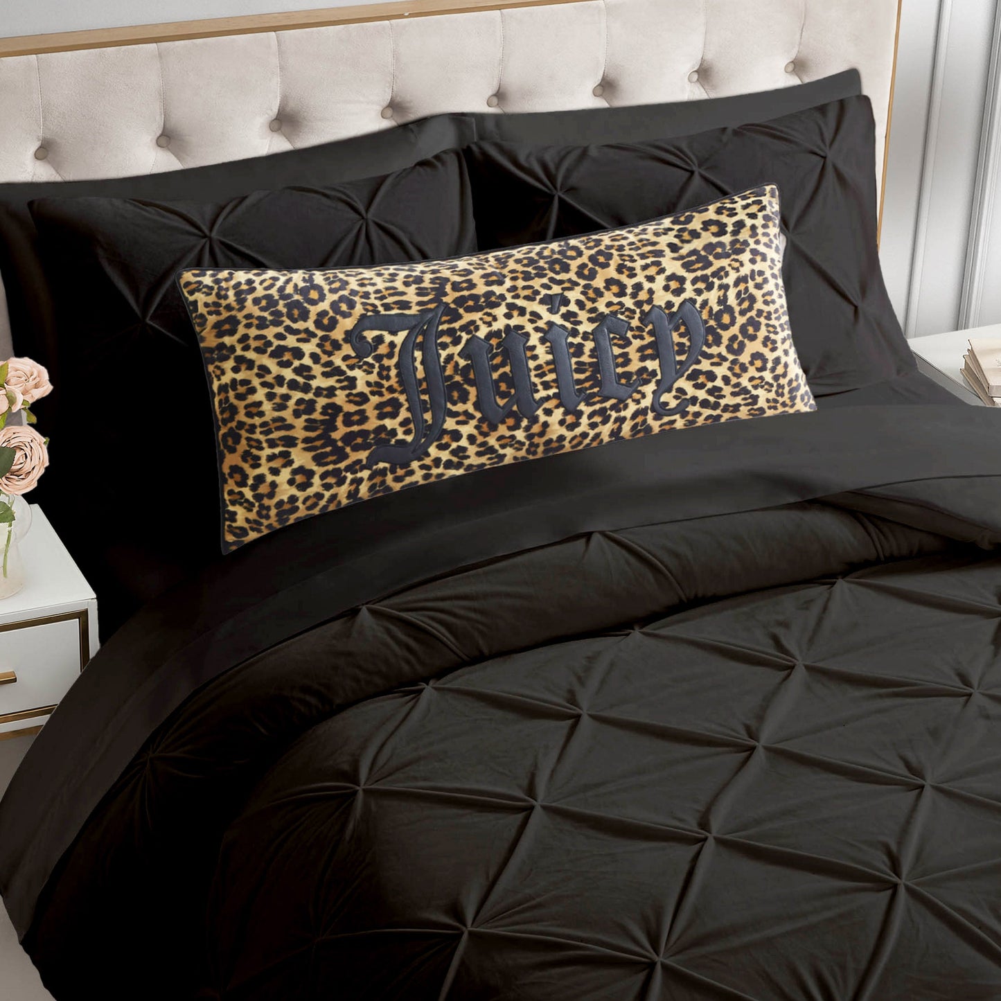 A bed with a tufted beige headboard, black bedding, and a rectangular Gothic Cheetah Decorative Pillow featuring leopard print and the word "Juicy" in bold black letters. A bedside table with pink roses is visible on the left.
