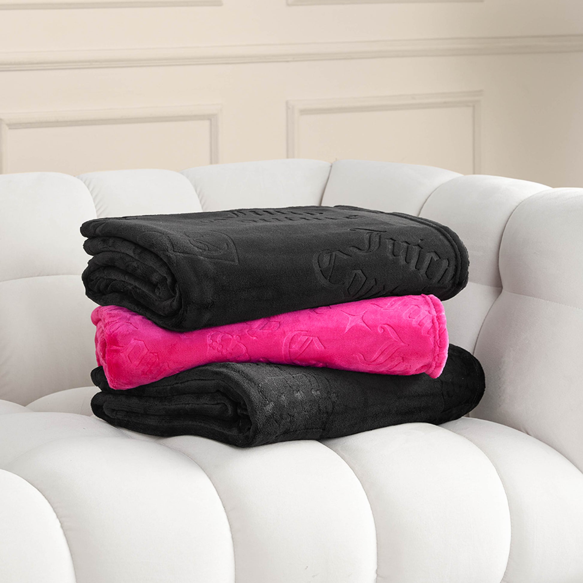 Three folded blankets—two black and one bright pink Embossed Plush Throw Blanket—are stacked on a white, tufted sofa in front of a beige wall with decorative molding.