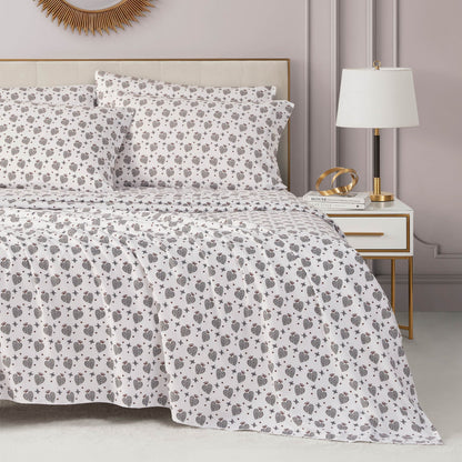 A stylish, light gray bedroom features a neatly made bed with a white and brown floral comforter, luxury pillows, and the Microfiber Sheet Set With Extra Pillowcases beside a white and gold nightstand holding books, gold decor, and a modern lamp.