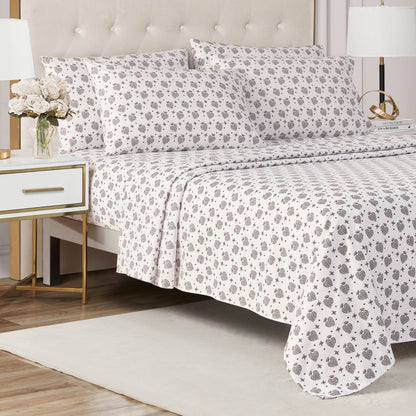 A neatly made bed with the Microfiber Sheet Set With Extra Pillowcases—white sheets and pillowcases adorned with a gray and black floral pattern—stands beside a white nightstand holding a vase of flowers in a bright, modern bedroom.