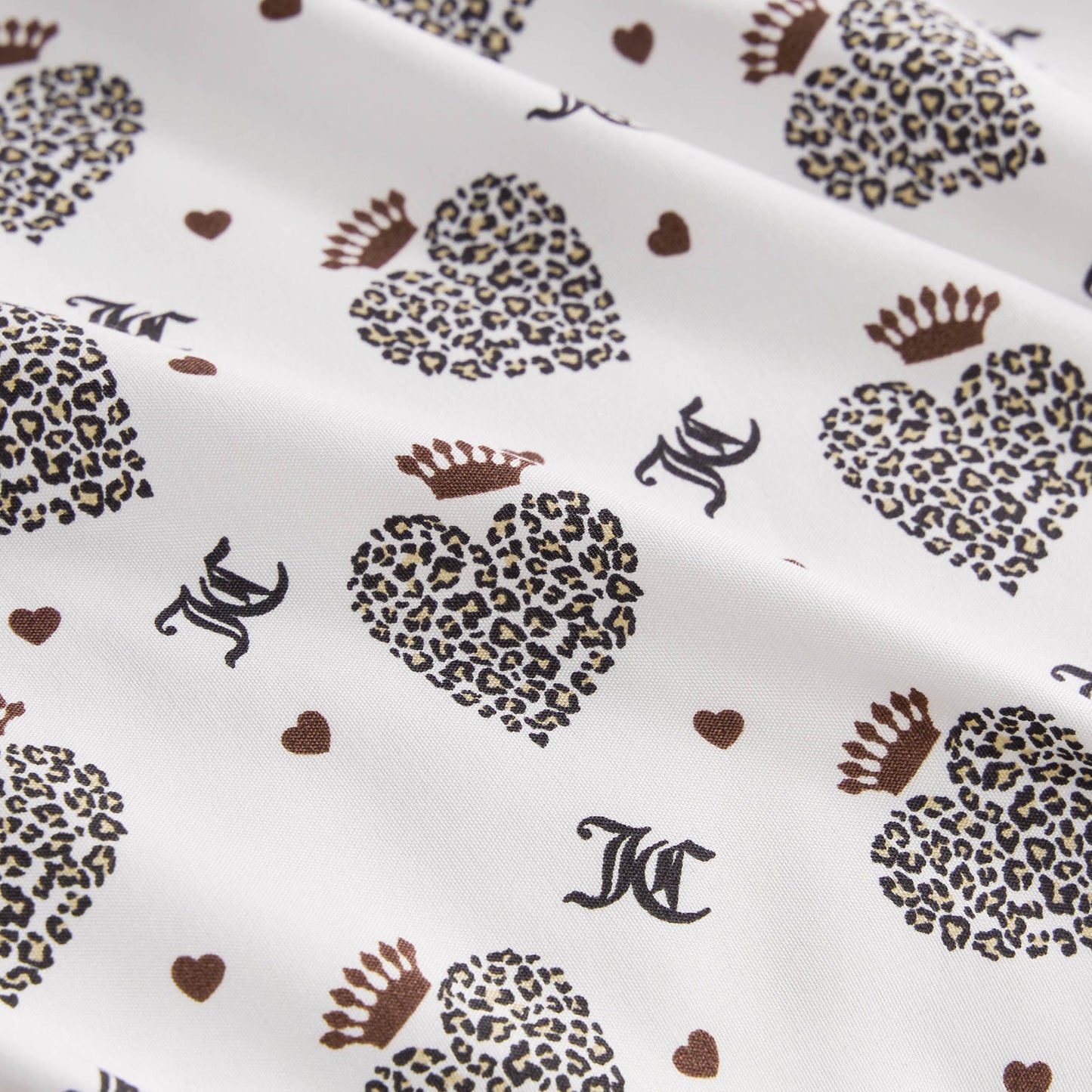 The Microfiber Sheet Set With Extra Pillowcases features white fabric with leopard print hearts wearing crowns, small brown hearts, and black JC letters, offering a soft, slightly wrinkled texture for luxurious bedding.