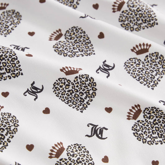 The Microfiber Sheet Set With Extra Pillowcases features white fabric with leopard print hearts wearing crowns, small brown hearts, and black JC letters, offering a soft, slightly wrinkled texture for luxurious bedding.