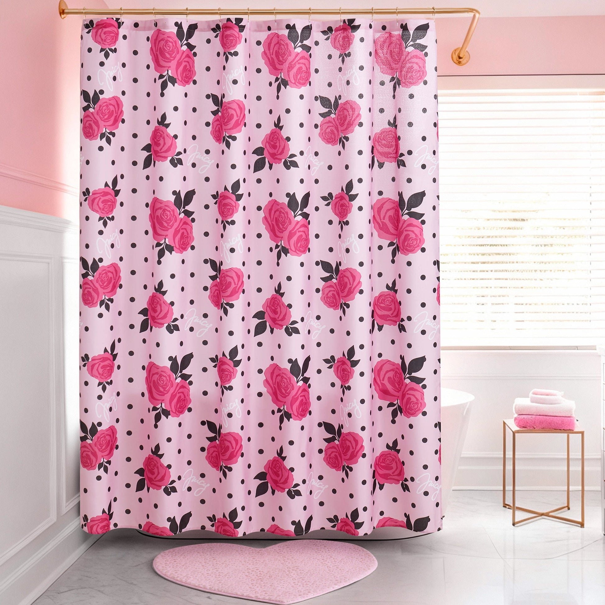A pink bathroom with a Shower Curtain and Hooks Set featuring red roses and black polka dots, a pink heart-shaped rug, white tub, folded towels on a stool, and a window with white blinds.