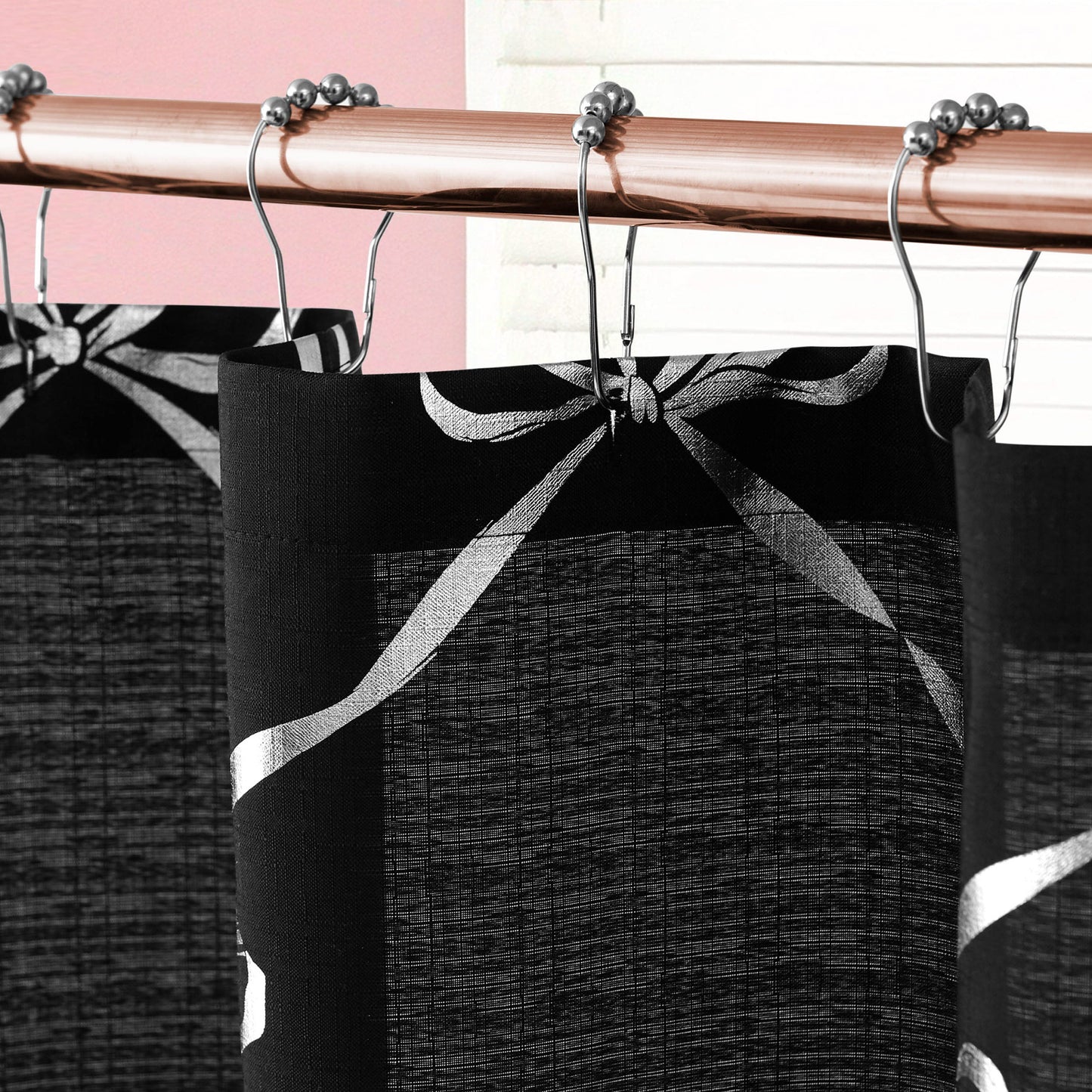 Close-up of a Shower Curtain and Hooks Set featuring a black and white bow pattern, hanging on a copper rod; chic bathroom decor is completed by pink and white walls.