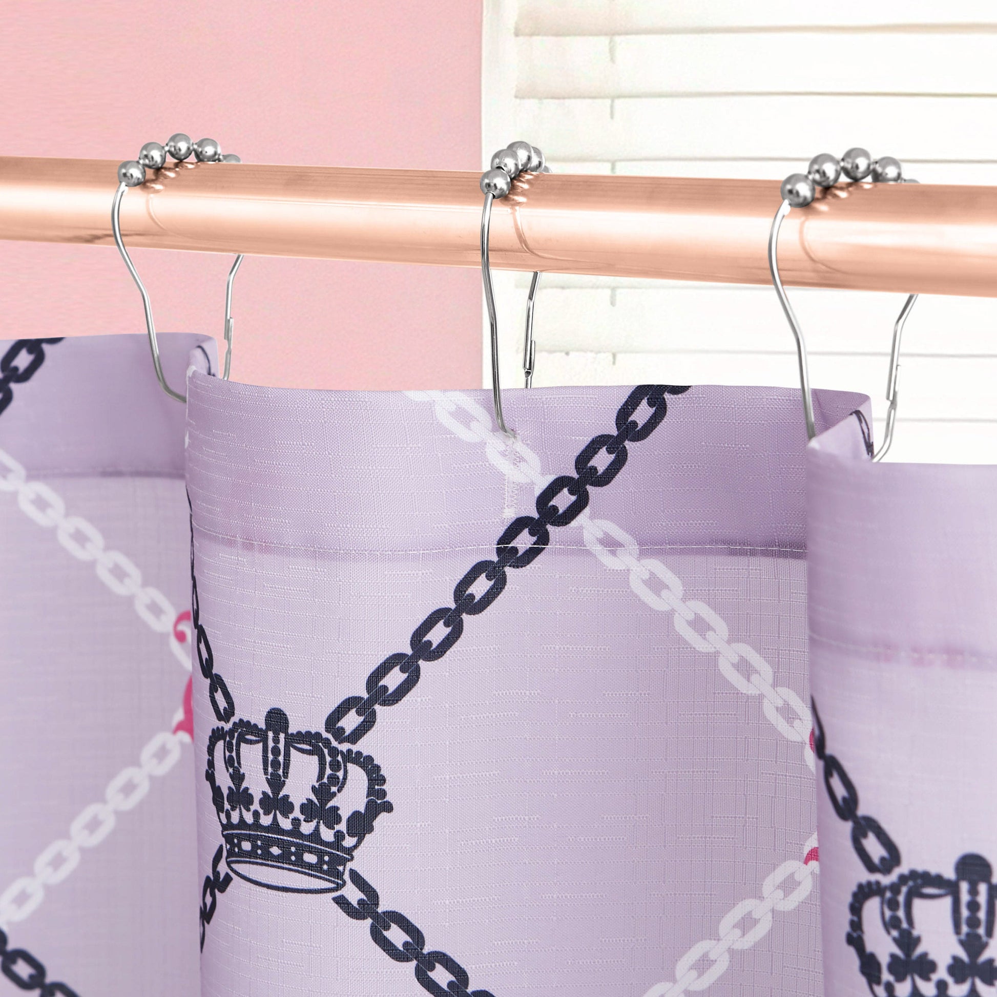 A close-up of the Shower Curtain and Hooks Set by Juicy Couture, showcasing a purple chain and crown pattern on a curtain hanging from metallic hooks. A white window blind and pink wall complement the chic bathroom decor.