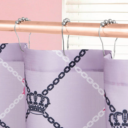 A close-up of the Shower Curtain and Hooks Set by Juicy Couture, showcasing a purple chain and crown pattern on a curtain hanging from metallic hooks. A white window blind and pink wall complement the chic bathroom decor.