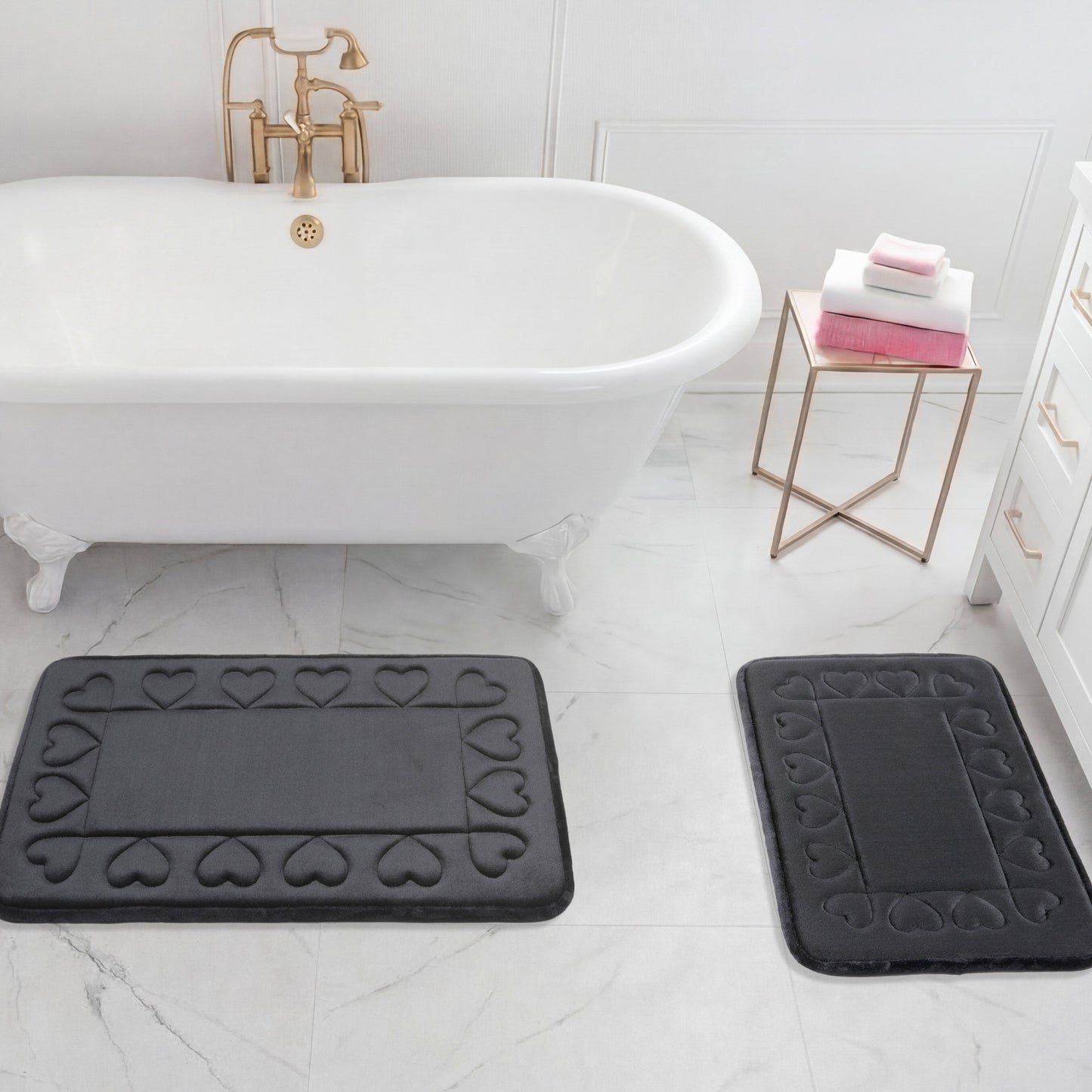A white clawfoot bathtub sits on a marble floor with two black Quilted Memory Foam Bath Mats in front. A side table holds folded towels beside white drawers with gold handles.