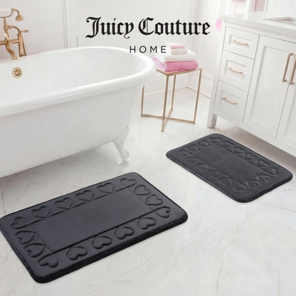 Two dark gray Quilted Memory Foam Bath Mats from Juicy Couture rest on a white marble floor near a clawfoot tub and vanity, adding luxury to the space. Juicy Couture Home is written at the top of the image.
