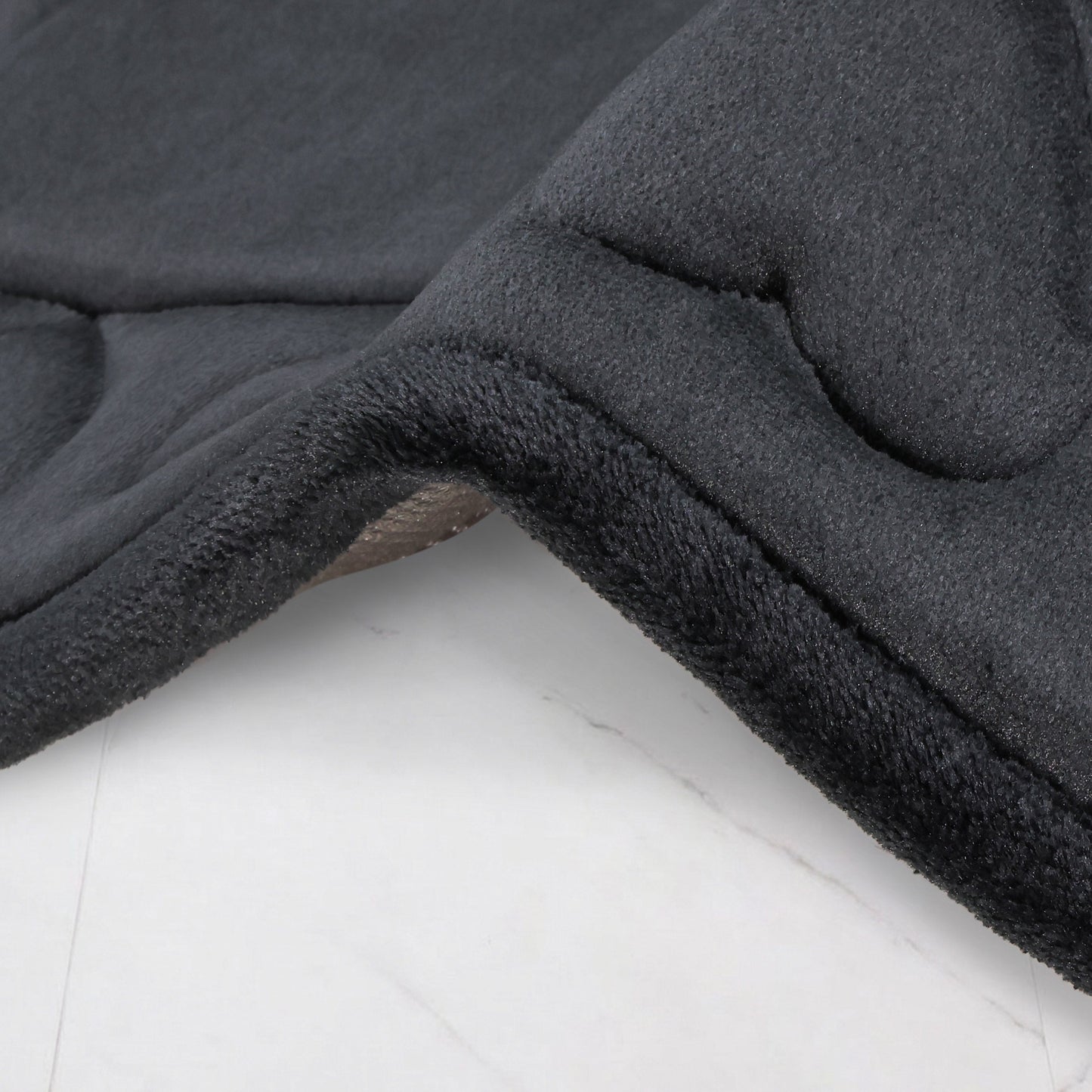 A close-up of a dark gray Quilted Memory Foam Bath Mat from a 2-piece set, draped over a light surface, highlighting its plush, soft texture and curved edge.