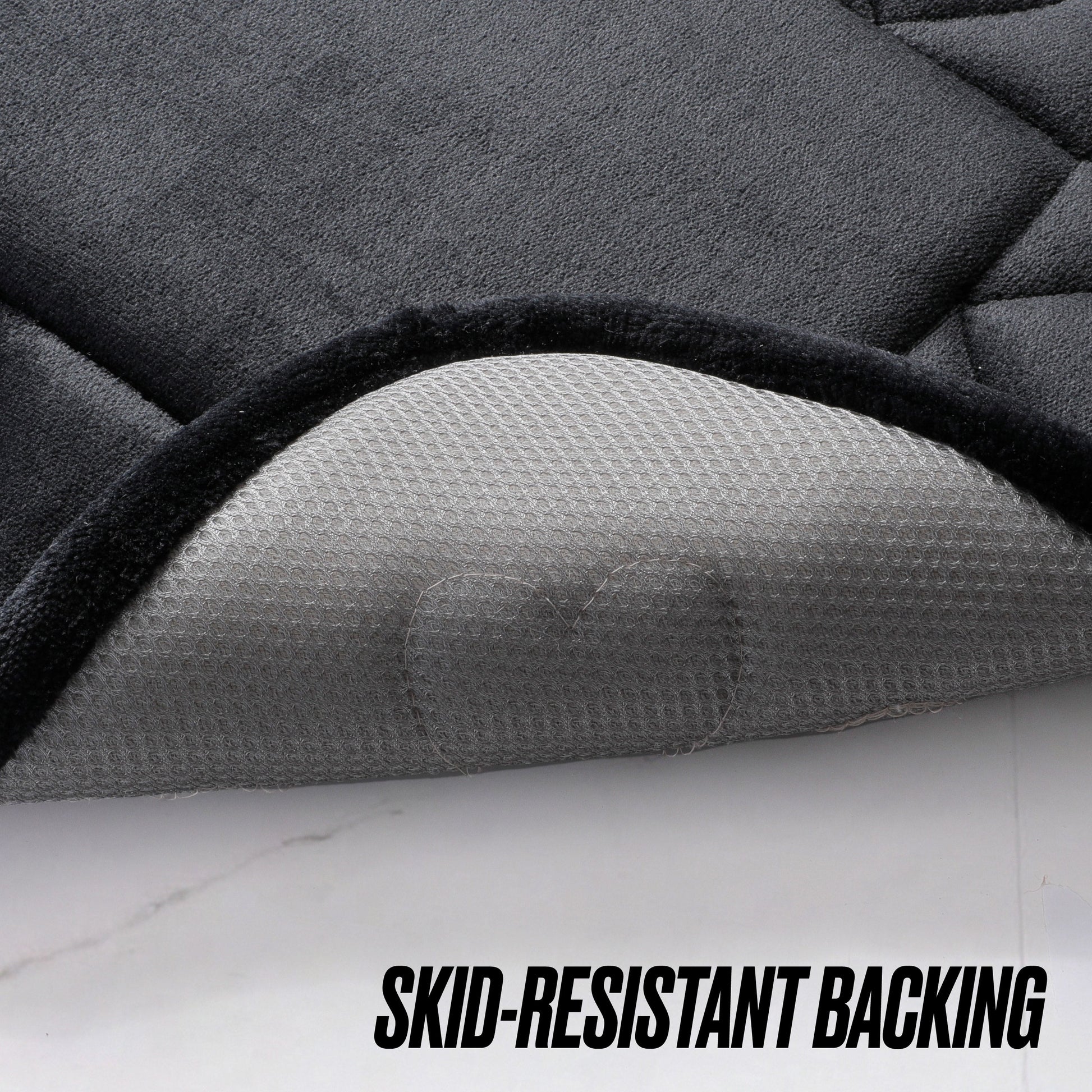 A close-up of a black Quilted Memory Foam Bath Mat 2-Piece Set with one corner lifted to reveal its gray, textured, skid-resistant backing. Bold black letters at the bottom read "SKID-RESISTANT BACKING.