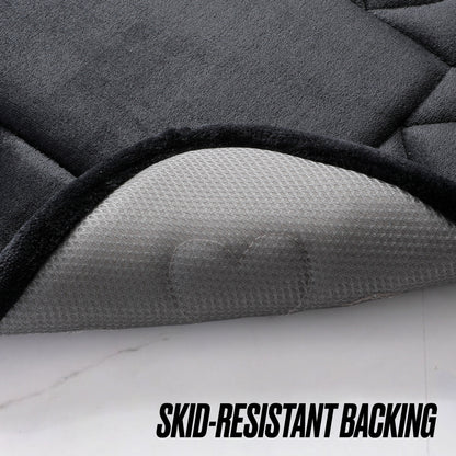 A close-up of a black Quilted Memory Foam Bath Mat 2-Piece Set with one corner lifted to reveal its gray, textured, skid-resistant backing. Bold black letters at the bottom read "SKID-RESISTANT BACKING.