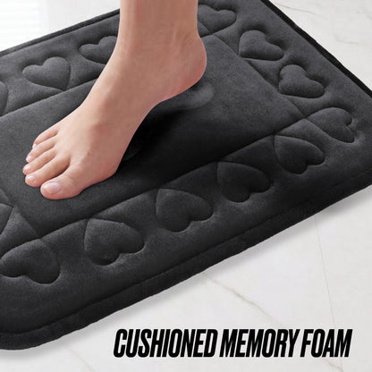 A bare foot steps onto the Quilted Memory Foam Bath Mat 2-Piece Set, featuring a black heart-patterned quilted design, placed on light tile flooring. Text at the bottom reads: CUSHIONED MEMORY FOAM.