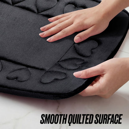 A person’s hands touch the edge of a black Quilted Memory Foam Bath Mat 2-Piece Set with quilted heart patterns on a white surface. Text at the bottom reads, SMOOTH QUILTED SURFACE.