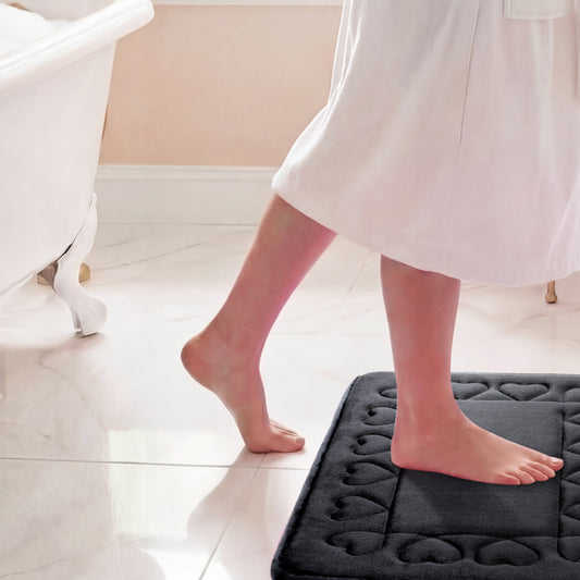 A person in a white robe steps barefoot onto the Quilted Memory Foam Bath Mat 2-Piece Set, bringing comfort and luxury to the bright, tiled bathroom.